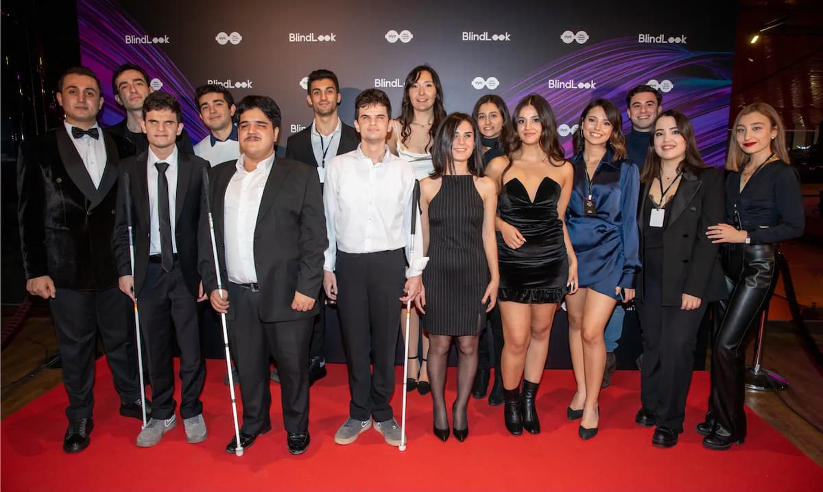 On a purple and black background on the screen, there is a white drawing with an eyeglasses icon inside it, and the text "EyeBrand." In front of the scene, the BlindLook team is smiling happily towards the cameras.